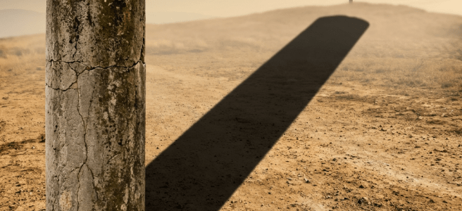Weathered stone column casting a long shadow toward a cross on a distant hill.