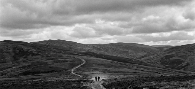 Wide moorland landscape with two hikers on a winding dirt path under cloudy sky