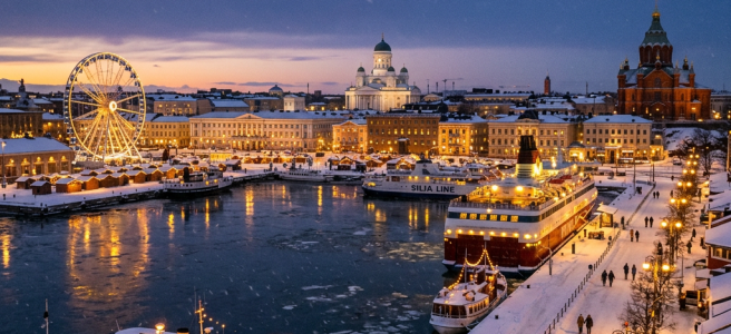 Helsinki harbor covered in snow with illuminated ferris wheel, boats, and historic buildings at dusk