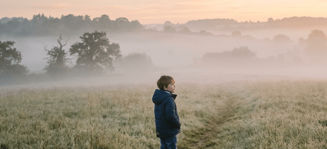Young boy in blue coat and boots standing on a grassy path in a misty field at dawn