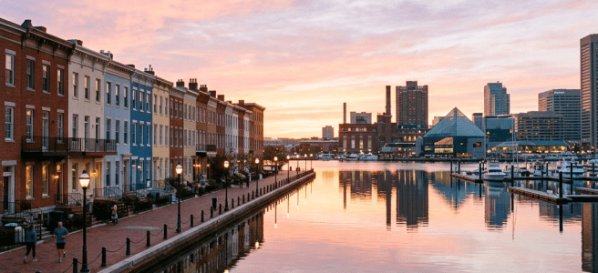 Colorful row houses along a harbor with calm water and city skyline at sunset